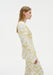 A woman stands in side profile against a plain white background, wearing a cream-colored long-sleeve top with a yellow floral pattern and matching trousers. Her straight light brown hair is worn down, and she maintains a neutral expression.
