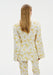 Rear view of a woman standing against a plain white background, wearing a cream-colored long-sleeve top with a yellow floral pattern and matching trousers. Her straight light brown hair falls down her back.