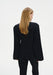 A woman with straight blonde hair is shown from the back in a relaxed standing pose against a plain background, her head slightly turned to the side.