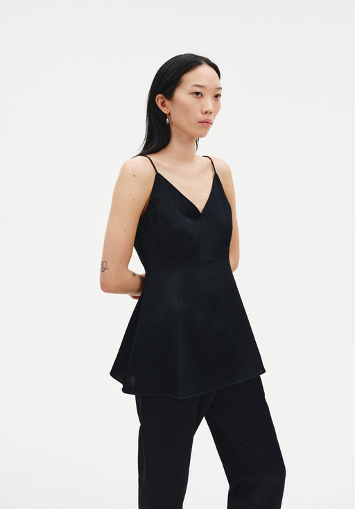 A woman stands in a three-quarter pose against a plain white background, wearing a black sleeveless top with thin straps and a flared hem, paired with black trousers. Her dark shoulder-length hair is worn down, and small tattoos are visible on her arms as she looks slightly to the side with a neutral expression.