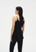 Rear three-quarter view of a woman against a plain white background, wearing a black sleeveless top with thin straps and black trousers. Her dark shoulder-length hair is worn down, and small tattoos are visible on her upper back and arms.