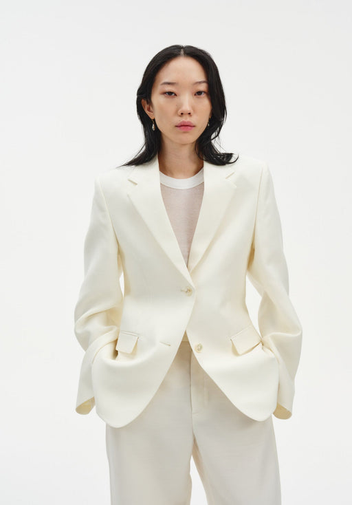 A woman faces forward against a plain white background, wearing a cream-colored tailored blazer with a single-button closure over a light top, paired with matching trousers. Her dark, shoulder-length hair is worn down, and she has a neutral expression with her hands in her pockets.