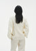 Rear view of a woman standing against a plain white background, wearing a cream-colored tailored blazer with structured shoulders and matching trousers. Her straight dark hair falls to mid-back, and her hands rest casually in her pockets.