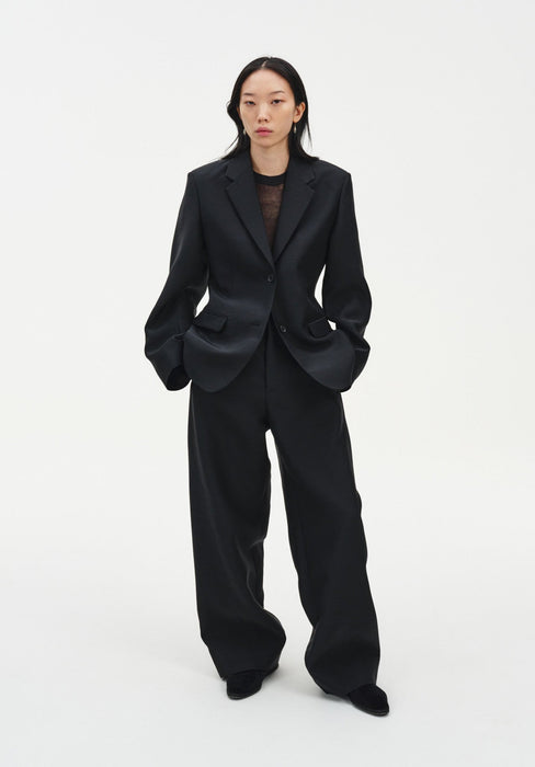 A woman stands facing forward against a plain white background, wearing a black tailored blazer over a sheer dark top with matching wide-leg trousers and black shoes. Her dark shoulder-length hair is worn down, and she maintains a neutral expression with her hands in her pockets.