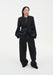 A woman stands facing forward against a plain white background, wearing a black tailored blazer over a sheer dark top with matching wide-leg trousers and black shoes. Her dark shoulder-length hair is worn down, and she has a neutral expression with her hands in her pockets.