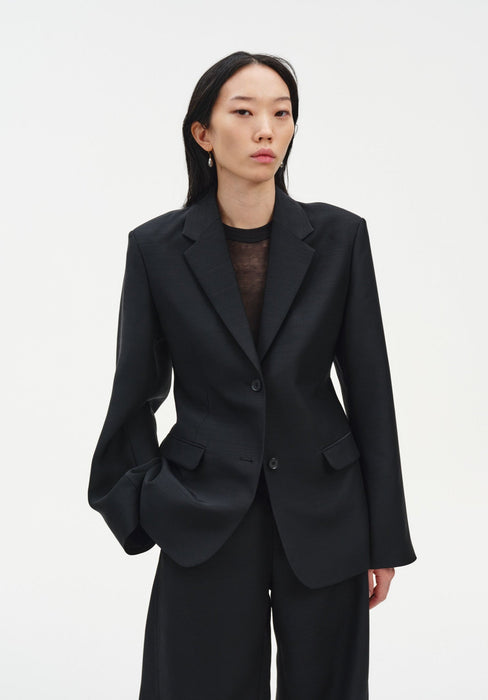 A woman stands facing forward against a plain white background, wearing a black tailored blazer over a sheer dark top with matching trousers. Her dark shoulder-length hair is worn down, and she has a neutral expression, with one hand in her pocket.