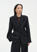 A woman stands facing forward against a plain white background, wearing a black tailored blazer over a sheer dark top with matching trousers. Her dark shoulder-length hair is worn down, and she has a neutral expression, with one hand in her pocket.