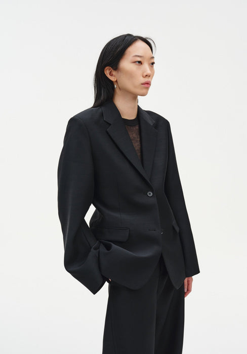 A woman stands in a three-quarter side pose against a plain white background, wearing a black tailored blazer over a dark top with matching trousers. Her dark shoulder-length hair is worn down, and she looks off to the side with a neutral expression, one hand in her pocket.
