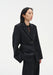 A woman stands in a three-quarter side pose against a plain white background, wearing a black tailored blazer over a dark top with matching trousers. Her dark shoulder-length hair is worn down, and she looks off to the side with a neutral expression, one hand in her pocket.