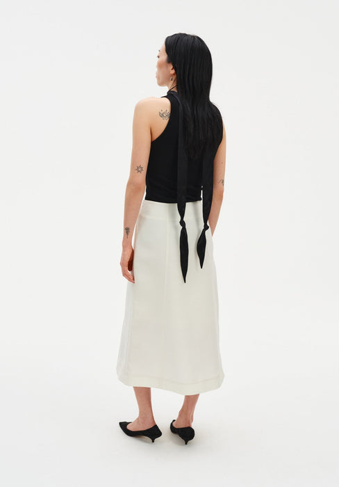 Rear three-quarter view of a woman against a plain white background, wearing a black sleeveless top with a long tie detail draping down the back, paired with a cream-colored midi skirt and black pointed heels. Her dark shoulder-length hair is worn down, and small tattoos are visible on her arms and upper back.