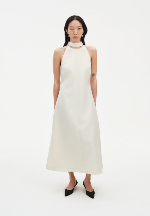 Front view of a model wearing a sleeveless cream midi dress with a high choker-style neckline and A-line silhouette, paired with black pointed shoes against a plain white background.