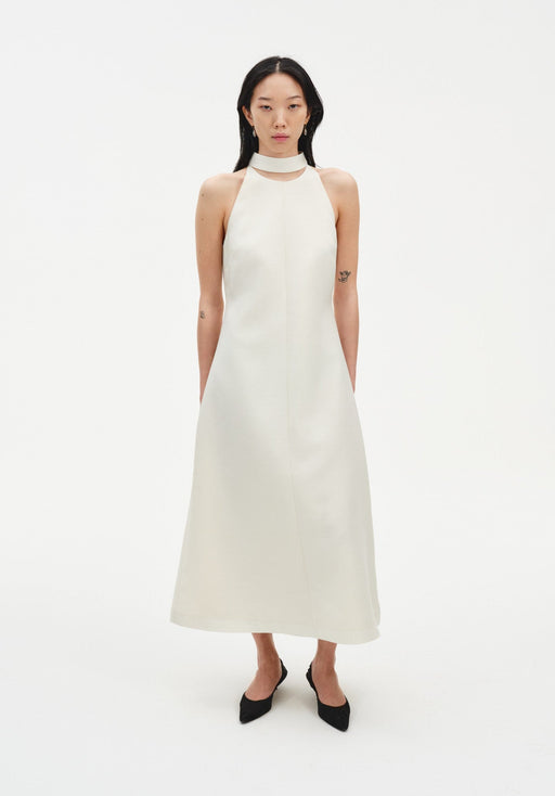 Front view of a model wearing a sleeveless cream midi dress with a high choker-style neckline and A-line silhouette, paired with black pointed shoes against a plain white background.