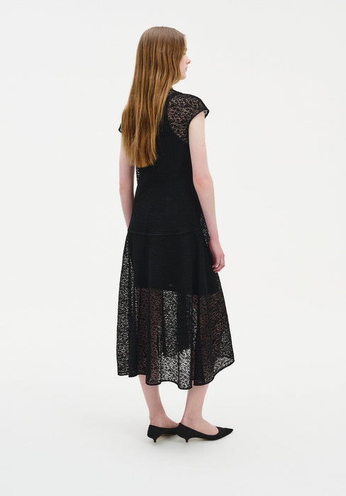 Three-quarter back view of a woman wearing a black semi-sheer lace midi dress with cap sleeves and a softly flared skirt, paired with black low heels in a minimal studio setting.