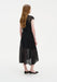 Three-quarter back view of a woman wearing a black semi-sheer lace midi dress with cap sleeves and a softly flared skirt, paired with black low heels in a minimal studio setting.