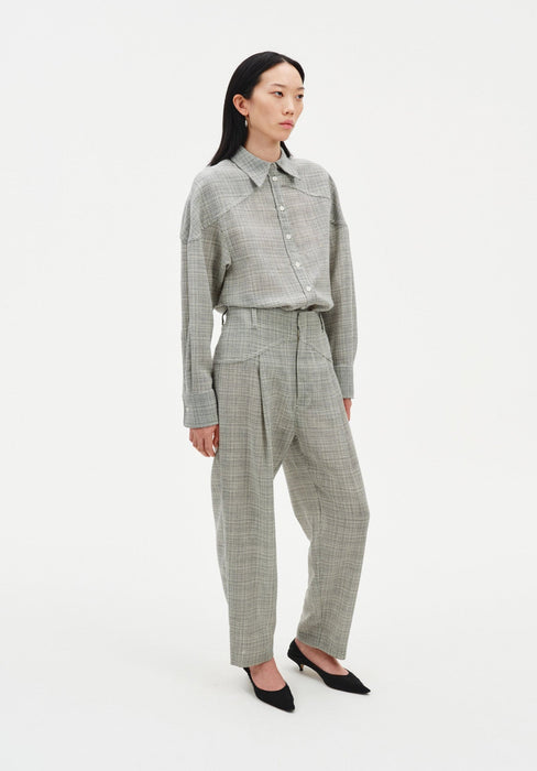 Full-length angled view of a woman wearing a grey textured button-up shirt tucked into matching high-waisted trousers, featuring a relaxed fit and subtle pleats, paired with black pointed heels against a clean studio background.