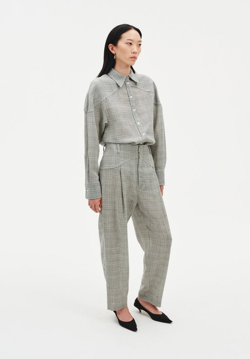 Full-length angled view of a woman wearing a grey textured button-up shirt tucked into matching high-waisted trousers, featuring a relaxed fit and subtle pleats, paired with black pointed heels against a clean studio background.