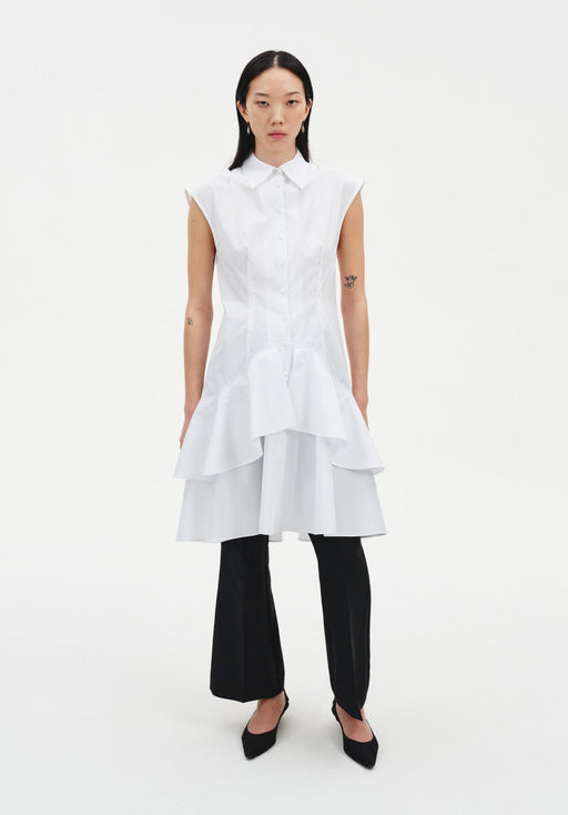 Front view of a model wearing a sleeveless white button-down tunic with a pointed collar and layered flared hem, styled over black slim trousers and paired with black pointed heels.
