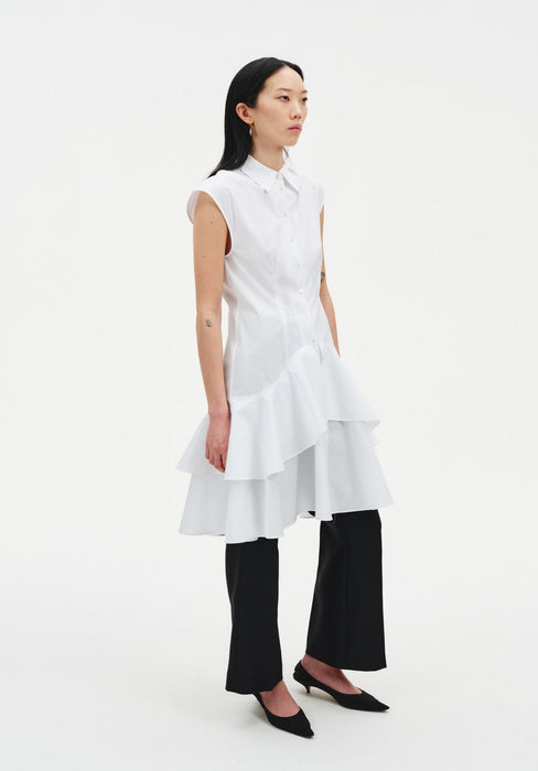 Side view of a model wearing a sleeveless white button-down tunic with a pointed collar and layered ruffled hem, styled over black cropped trousers and paired with black pointed heels.
