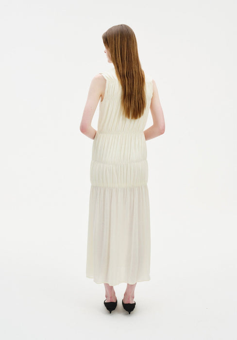 Rear view of a woman standing against a plain white background, wearing a sleeveless cream-colored midi dress with gathered tiers and a softly flowing skirt. Her straight light brown hair falls down her back, and she is wearing black pointed heels.