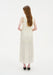 Rear view of a woman standing against a plain white background, wearing a sleeveless cream-colored midi dress with gathered tiers and a softly flowing skirt. Her straight light brown hair falls down her back, and she is wearing black pointed heels.